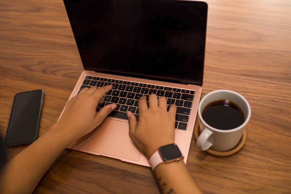 Stratégie de communication inclusive. Une personne écrit sur un clavier d'ordinateur portable. On ne voit que ses avant-bras et ses mains sur le clavier. À gauche de l'ordinateur, un téléphone portable, éteint. À droite, une tasse de café.
